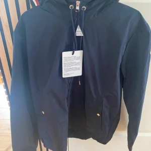 Mörkblå vindjacka från Moncler - Snygg mörkblå vindjacka från Moncler med huva och dragkedja framtill. Jackan har guldiga detaljer, snörning i huvan och Moncler-logga på ärmen. Materialet är lätt och känns sportigt, perfekt för blåsiga dagar. Fodret i huvan har en cool randig detalj i rött, vitt och orange.