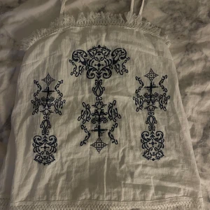 Vit linnetopp med blå broderi - Snygg vit linnetopp med tunna axelband och dekorativa blå broderier framtill. Toppen har fransdetaljer upptill och spetskant nertill för en bohemisk vibe. Perfekt för varma dagar och festivaler. Storlek L men sitter mer som en M/S. Aldrig använd 