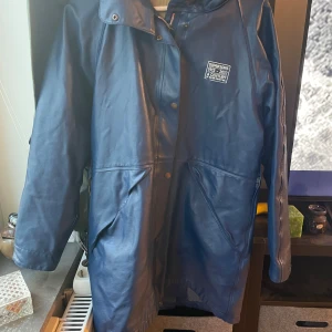 Blå regnjacka från Didriksons - Säljer en blå regnjacka från Didriksons med huva och tryckknappar framtill. Jackan har två stora fickor och ett diskret tryck på bröstet. Perfekt för regniga dagar och har en klassisk, enkel design som funkar till allt.