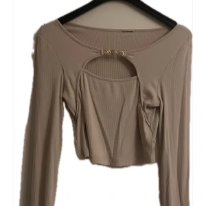 Beige ribbad croppad långärmad topp - Trendig beige croppad topp med långa ärmar och ribbad struktur. Snygg cut-out detalj framtill med guldfärgat spänne. Använd bara en gång och tog bort tagen bak då den kliade plagget är från hemsidan Dazy.