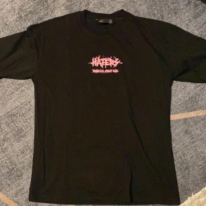 Svart t-shirt med tryck från Bershka - Svart t-shirt från Bershka med stort grafiskt tryck i rosa och vitt på ryggen och mindre tryck framtill. Trycket har street-inspirerad design med texten 'HATERS' och en maskerad figur. Klassisk passform och rund hals. Perfekt för dig som gillar edgy streetstyle.