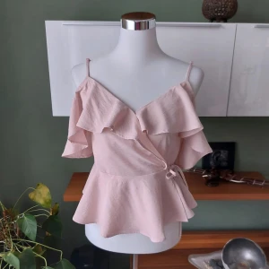 Ljusrosa omlottopp med volang - Supersöt ljusrosa omlottopp med volangdetaljer och smala axelband. Toppen har korta ärmar och knyt i sidan för en feminin look. Perfekt för dig som gillar romantisk och trendig stil.