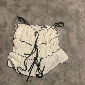 Vit linnetopp med svarta detaljer Gina Tricot - Supersöt vit linnetopp från Gina Tricot med svarta kontrastsömmar och knytband framtill. Toppen har tunna axelband och volangiga lager som ger en fin och elegant look. Perfekt för dig som gillar trendiga och unika toppar med extra detaljer.