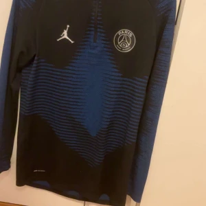 PSG långärmad träningströja Jordan - Snygg långärmad fotbollströja från PSG och Jordan. Tröjan är svart med blått mönster och har PSG-logga samt Jumpman-logga på bröstet. Tillverkad i ett tekniskt material som andas, perfekt för träning eller match.