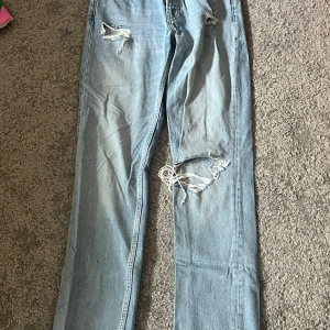 Ljusblå slitna jeans med hål lågmidjade  - Säljer ett par ljusblå jeans med flera slitningar och hål på benen för en riktigt avslappnad och trendig look. Jeansen har rak passform och klassisk femficksdesign. Perfekta för dig som gillar en edgy streetstil. Hål i fickan då det var ett rött tyg i hålen som jag klippt bort 