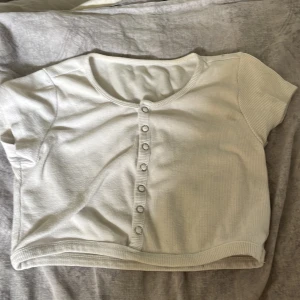 Vit ribbad croppad topp med knappar - Säljer en vit, croppad topp med korta ärmar och ribbad struktur. Toppen har rund halsringning och en rad med knappar framtill som ger en chill och trendig vibe. Perfekt att matcha med jeans eller kjol för en enkel men snygg look.