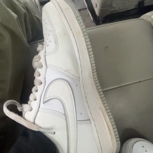 Nike Air Force 1 vita sneakers - Klassiska Nike Air Force 1 sneakers i helvitt skinn med perforerad tå, rund tå och platt sula. Snygg low-top modell med vita snören och ikonisk Swoosh-logga på sidan. Perfekta för en clean och stilren look.
