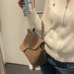 Brun handväska i skinn med unik form - Säljer en stilren brun handväska i skinn med ett snyggt, asymmetriskt lock och kort handtag. Väskan har en minimalistisk design som passar perfekt till jeans eller en enkel outfit. Rymmer det viktigaste och är lätt att bära med sig.