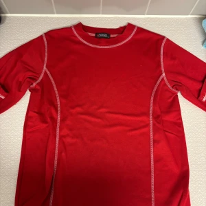 Röd långärmad tröja från Rogue - Snygg röd långärmad tröja från Rogue med kontrastsömmar i vitt. Tröjan har en klassisk rund halsringning och är tillverkad i mjuk bomull, perfekt för en avslappnad och sportig stil.