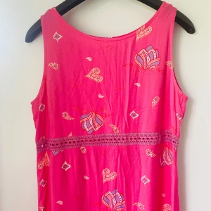 Lång rosa klänning med blommigt mönster - Superfin vintage långklänning i stark rosa med blommigt och paisley-inspirerat mönster i lila, vitt och rött. Klänningen är ärmlös, har en markerad midja med spetsdetalj och dragkedja bak. Perfekt för sommarens alla tillfällen! Märkt storlek 40 men passar perfekt för 36/38