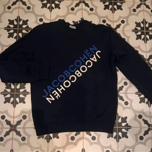 Mörkblå sweatshirt från Jacob Cohën - Snygg mörkblå sweatshirt från Jacob Cohën med stort tryck i blått och vitt diagonalt över bröstet. Tröjan har rund halsringning, långa ärmar och ribbade muddar. Perfekt för en avslappnad och trendig stil. Tröjan är i nyskick utan skador eller defekter, hör av er vid funderingar!