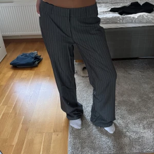 Randiga kostymbyxor från Madlady - Snygga mörkgrå kostymbyxor från Madlady med tunna vita ränder. Byxorna har raka ben, bälteshällor och klassisk passform. Perfekta för dig som gillar en stilren och trendig look. Materialet känns mjukt och bekvämt. Säljer pga för stora, aldrig använda. Helt nya.