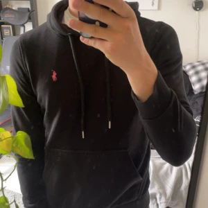 Svart hoodie från Polo Ralph Lauren - Svart hoodie från Polo Ralph Lauren med klassisk rosa broderad logga på bröstet. Tröjan har huva med snörning, magficka och ribbade muddar. Perfekt för en chill och stilren look.
