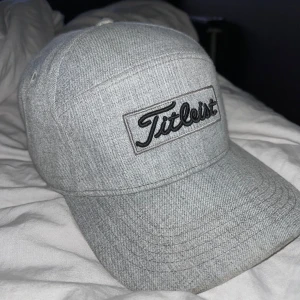 Grå Titleist keps i bomull - Snygg grå keps från Titleist med broderad logga framtill. Kepsen har klassisk böjd skärm, justerbar rem baktill och är tillverkad i mjuk bomull. Perfekt för golfbanan eller en chill dag ute.