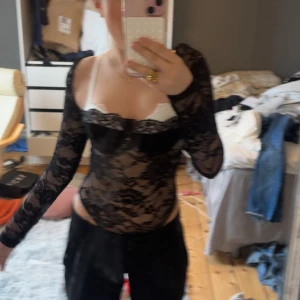 Svart spetsig body - Säljer en svart långärmad body i genomskinlig spets med figurnära passform. Toppen har v-ringad urringning och är perfekt för dig som gillar en edgy och trendig stil. Snygg att styla med jeans eller kjol för en cool look.