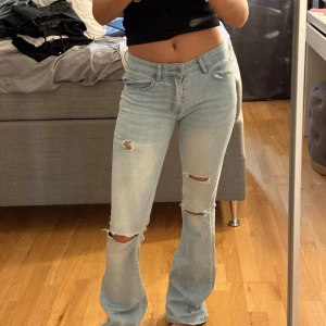 Ljusblå bootcut jeans med slitningar - Säljer ett par ljusblå jeans med bootcut passform och snygga slitningar på benen. Jeansen har klassisk femficksdesign, låg midja och är tillverkade i mjukt denimtyg. Perfekta för en avslappnad och trendig look. fläcken på bena går nog bort i tvätten