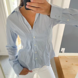 Ljusblå randig skjorta från H&M - Snygg ljusblå skjorta från H&M med tunna vita ränder. Klassisk krage, långa ärmar och knäppning framtill med vita knappar. Skjortan är i bomull och har en avslappnad passform som funkar till många olika stilar.