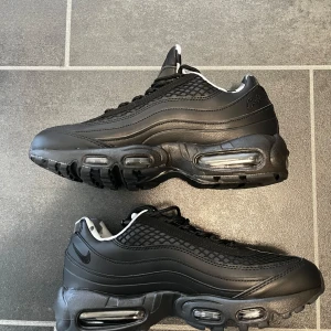 Nike Cortez Air Max 95 Svarta Sneakers - Svarta Nike Air Max 95 sneakers med coola detaljer och vågformade paneler i skinn och syntet. Ovandelen har mesh med rutmönster och snörning. Fodret har svartvit camo. Synliga Air-enheter i sulan ger en sportig vibe. Perfekta för dig som gillar streetstyle.