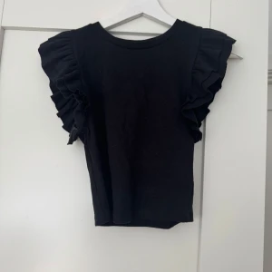 Svart volangtopp från Zara - Supersnygg svart kort topp från Zara med volangdetaljer. Toppen är i storlek M, men den är mer som en S.