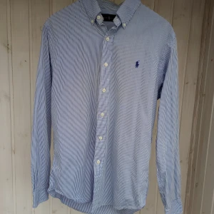 Randig skjorta från Ralph Lauren M - Snygg blå- och vitrandig skjorta från Ralph Lauren i klassisk modell. Skjortan har lång ärm, knappar framtill och ikonisk broderad logga på bröstet. Tillverkad i mjuk bomull och passar perfekt till jeans eller chinos. 