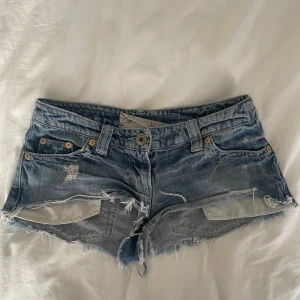 River Island slitna jeansshorts - Snygga blå jeansshorts från River Island med slitna detaljer och fransig kant. Klassisk femficksmodell med låg midja och guldiga nitar. Perfekta för sommaren när du vill ha en avslappnad och cool look.