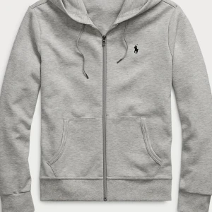 Grå hoodie från Polo Ralph Lauren - Snygg ljusgrå hoodie från Polo Ralph Lauren med dragkedja, huva och snörning. Klassisk logga broderad på bröstet och två stora fickor framtill. Perfekt för en chill och stilren look.