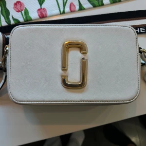 Vit axelväska Marc Jacobs Snapshot - Säljer en snygg vit axelväska från Marc Jacobs Snapshot med guldiga detaljer och bred, justerbar axelrem med logga. Väskan har två dragkedjor och är tillverkad i tåligt skinn med struktur. Perfekt för dig som vill ha en stilren och trendig accessoar.
