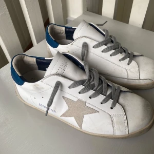 Golden Goose Superstar vita sneakers - Golden Goose Superstar sneakers i vitt skinn med grå mockastjärna på sidan, grå snörning och blå detaljer vid hälen. Klassisk rund tå och platt sula. Snyggt slitna detaljer för en cool look. Perfekta för dig som gillar streetstyle.