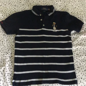 Randig pikétröja Polo Ralph Lauren XS - Mörkblå pikétröja från Polo Ralph Lauren i slim fit, med vita horisontella ränder och broderad nalle på bröstet. Klassisk krage med knappar och korta ärmar. Perfekt för dig som har bra stil och snygga detaljer. På sista bilden var det spegeln som var smutsig och inte piken 