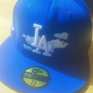Blå LA Dodgers keps från New Era - Säljer en blå 59FIFTY keps från New Era med LA Dodgers brodyr framtill och molndetaljer. Kepsen har broderad text på sidan och MLB-logga bak. Klassisk flat peak och strukturerad passform. Perfekt för dig som vill sticka ut med sportig stil.