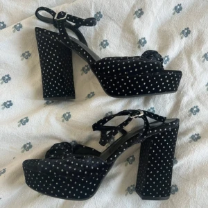 Svarta platåsandaler med prickar M&S - Aldrig använt. Ny med lappen kvar. Snygga svarta velvet sandaler från M&S med höga blockklackar och platåsula. Skorna har ett prickt mönster i silver, knytdetalj framtill och justerbar rem runt vristen. Tillverkade i mjuk sammet för en lyxig känsla.