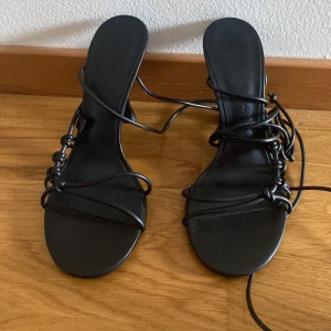 Svarta sandaletter med snörning från Mango - Snygga svarta sandaletter från Mango med tunna remmar och snörning runt foten. Skorna har öppen tå, låg klack och stilren design i syntetmaterial. Perfekta för att lyfta din outfit med en trendig touch.