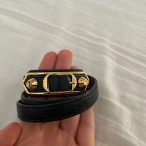 Svart Balenciaga armband vintage med gulddetaljer - Snyggt svart armband från Balenciaga i äkta skinn med exklusiva guldfärgade metalldetaljer och spänne. Armbanet är vintage. Perfekt accessoar för att lyfta din outfit med lyxig känsla.