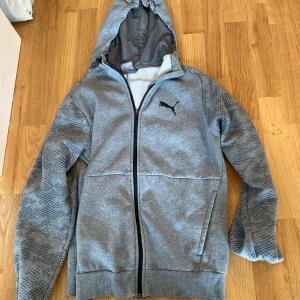 Grå hoodiejacka från Puma - Grå hoodiejacka från Puma med svart logga på bröstet. Jackan har hel dragkedja, huva och diskret mönster på ärmarna. Tillverkad i mjukt material, perfekt för chill eller träning. Snygg och sportig look med ribbade muddar och praktiska fickor.
