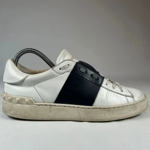 Valentino Garavani Open sneakers vit/svart - Snygga Valentino Garavani Open sneakers i vitt skinn med bred svart läderdetalj över mitten. Klassisk rund tå, platt sula och coola nitar baktill på hälen. Perfekta för dig som gillar stilrena men ändå unika sneakers.