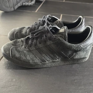 Svarta adidas Gazelle i mocka - Säljer ett par svarta adidas Gazelle med klassiska tre ränder på sidan. Skorna är i mocka med detaljer i skinn och har snörning. Snygg och tidlös modell med platt sula och rund tå, perfekt för dig som gillar streetstyle. Passar dig som bär storlek 38/39