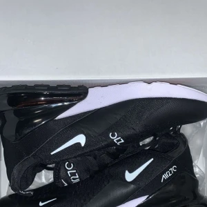 Svarta Nike Air Max 270 sneakers - Nike Air Max 270 sneakers i svart med vita detaljer och logga. Skorna har en stor synlig Air-enhet i hälen, mesh-överdel och snörning. Snygg och sportig design med bekväm passform och platt sula. Perfekta för dig som gillar streetstyle och sportiga vibes. Storlek 43