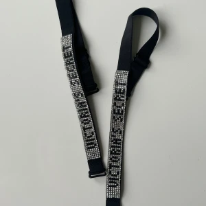 Svart bh band? - Snyggt svart bälte med justerbar längd och glittriga silverfärgade stenar längs framsidan. Perfekt accessoar för att lyfta din outfit med extra bling. Bältet har en smal och stilren design med metallspänne och elastiskt band.