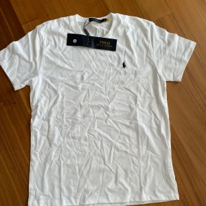 Vit Polo Ralph Lauren T-shirt - XL - *   Polo Ralph Lauren T-shirt *   Storlek: XL *   Vit med marinblå logga *   Helt ny – aldrig använd  Stilren och oanvänd T-shirt från Polo Ralph Lauren i storlek XL. Perfekt för en avslappnad och sofistikerad stil. Den klassiska marinblå loggan är broderad på bröstet. Levereras med alla originaletiketter. Ett utmärkt tillskott till din garderob för en tidlös look! Kontakta mig om ni vill ha mer bilder