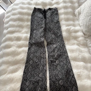 Grå ormmönstrade byxor från Monki - Säljer ett par grå byxor från Monki med ormmönster och snörning framtill. Byxorna har utsvängda ben och en cool, edgy vibe. Perfekta för dig som vill sticka ut med ett djurmönster och gillar trendiga detaljer.