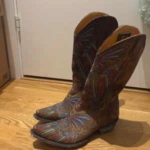 Broderade cowboy boots från Old Gringo - Unika cowboy boots från Old Gringo i brunt skinn med färgglada broderier i blått, rött och gult. Boots med spetsig tå och klassisk hög skaft, handgjorda i Mexiko. Inköps pris i USA runt $600-$700
