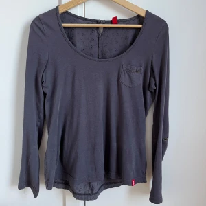 Grålila långärmad topp med spetsdetaljer - Supersnygg mörkgrå/lila långärmad topp från Esprit med rund halsringning, liten bröstficka och broderade spetsdetaljer på ryggen. Mjuk bomullskänsla och knapp i nacken och armarna. Perfekt för dig som gillar stilrena plagg med en twist. 💜