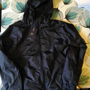 Svart Arsenal vindjacka med huva - Svart vindjacka från Arsenal med huva och dragkedja framtill. Jackan är tillverkad i lätt polyester och har en clean look utan mönster. Perfekt för dig som vill visa ditt stöd för Arsenal även när det blåser ute. #arsenal #fotboll