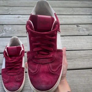 Valentino Garavani röda sneakers mocka - Snygga sneakers från Valentino Garavani i röd mocka med vita detaljer och coola nitar runt sulan. Skorna har snörning, rund tå och en vit platt sula med mönstrad undersida. Perfekta för dig som vill sticka ut med exklusiva och trendiga skor.