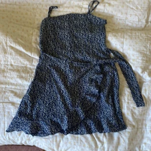 Blommig blå kortklänning från SHEIN - Supersöt blå kortklänning från SHEIN med vita blommor, smala axelband och volangkant nertill. Omlottknytning i midjan ger en snygg passform. Perfekt för sommarens alla tillfällen!