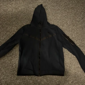 Svart Nike vindjacka med huva - Marinblå tech fleece från Nike med hel dragkedja och huva. Jackan har diskreta mörka ränder över bröstet och är tillverkad i ett lätt syntetmaterial. Perfekt för dig som gillar sportig stil och vill ha något enkelt men snyggt att slänga på dig.