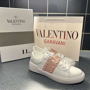 Valentino Garavani Rockstud sneakers - Snygga vita Valentino Garavani Rockstud sneakers med ljusrosa band och ikoniska nitar i guldton. Skorna har rund tå, platt sula och klassisk snörning. Tillverkade i skinn för en lyxig känsla och cool look. Perfekta för dig som vill sticka ut med stil.