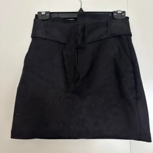 Svart mockakjol med bälte från Zara - Svart kortkjol i mocka från Zara med brett bälte och spänne i midjan. Kjolen har en stilren och enkel design som ger en snygg siluett. Perfekt för dig som gillar klassiska plagg med en twist.