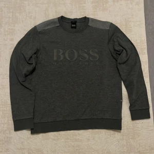 Grå slim fit tröja från Hugo Boss - Mörkgrå tröja från Hugo Boss med diskret logga framtill. Slim fit-modell med rund halsringning och ribbade muddar. Tröjan har en snygg kontrastpanel över axlarna och är tillverkad i mjuk bomull. Perfekt för en clean och stilren look.