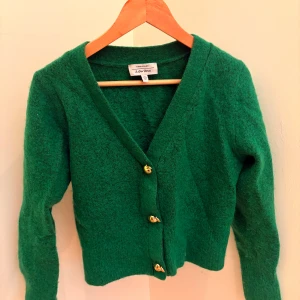 Grön kofta från & Other Stories - Superfin grön kofta från & Other Stories med unika guldfärgade knappar formade som små äpplen. Koftan är stickad, har v-ringning och långa ärmar. Perfekt för lager-på-lager och ger en färgklick till din outfit.